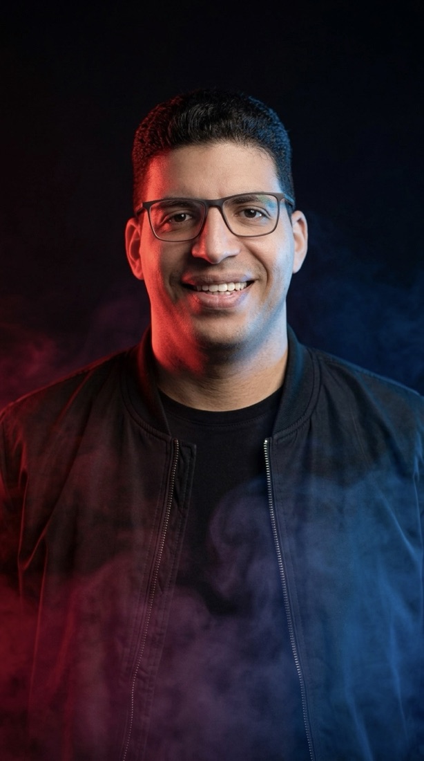 Zak smiling, in a black jacket, lit in red and blue